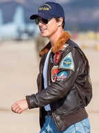 Person wearing a leather jacket with patches and a cap, standing on an airfield.