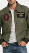 A man wearing an olive green bomber jacket with military-style patches on the sleeves and chest, layered over a white t-shirt and blue jeans. The jacket features a high collar, zippered front, and detailed embroidered graphics.