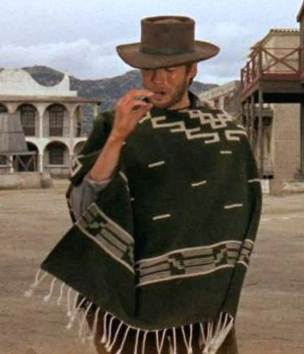 A man wearing a brown cowboy hat and a dark green patterned fringed poncho stands in a dusty western town street.