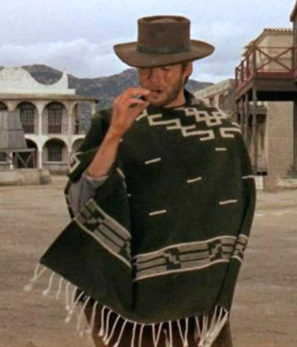 A man wearing a brown cowboy hat and a dark green patterned fringed poncho stands in a dusty western town street.