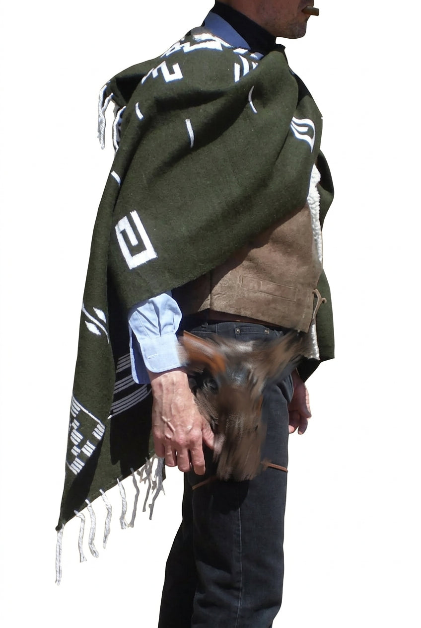 A man shown from the side wearing a dark olive green woven poncho with white geometric patterns and fringed edges over a brown vest, blue shirt, and black pants, with a brown holster containing a revolver at his hip.