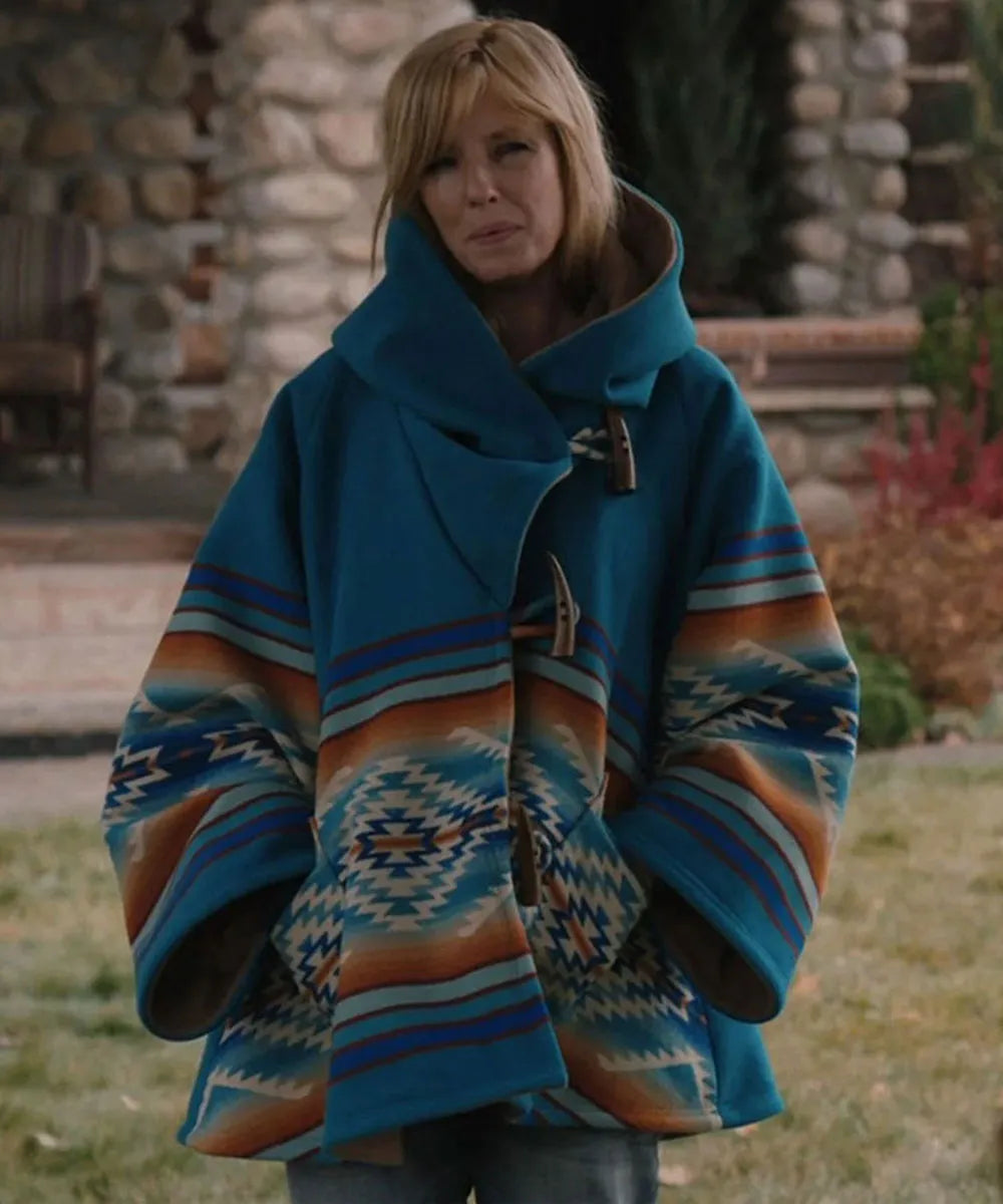 A woman wearing a vibrant blue and multicolor Southwestern-style patterned poncho with a high collar and toggle closures, standing outdoors in front of a stone wall.