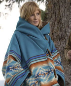 A woman wearing a blue hooded wrap poncho with Southwestern geometric patterns in orange, brown, and white, standing outdoors next to a tree.